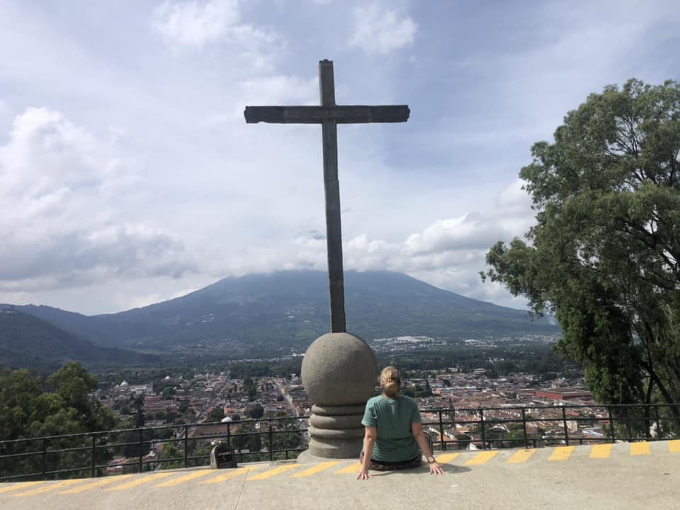 Person sitting on a ledge with a large cross in the background, overlooking a city and mountains.