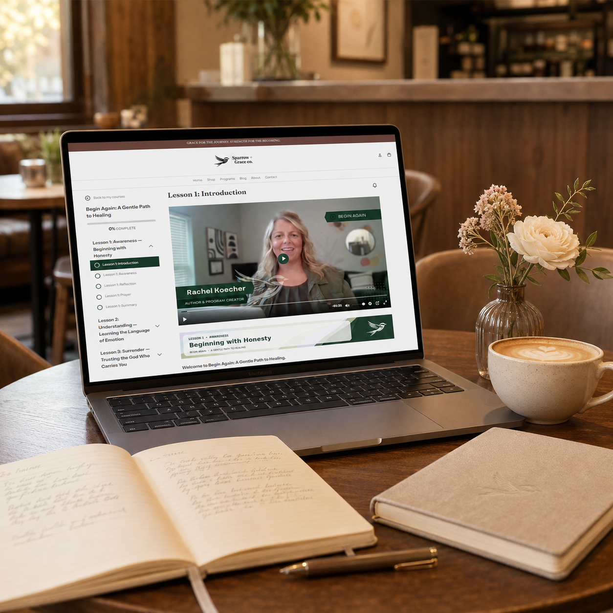 A laptop on a wooden table in a cafe setting, displaying a video lesson page. In front of the laptop are an open notebook, a pen, a closed notebook, and a cup of coffee, with a small vase of flowers to the right.