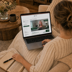 A woman sits on a beige couch in a cozy living room, viewed from behind, using a laptop to watch a video course. She is wearing a beige ribbed sweater and has an open notebook and pen on her lap.