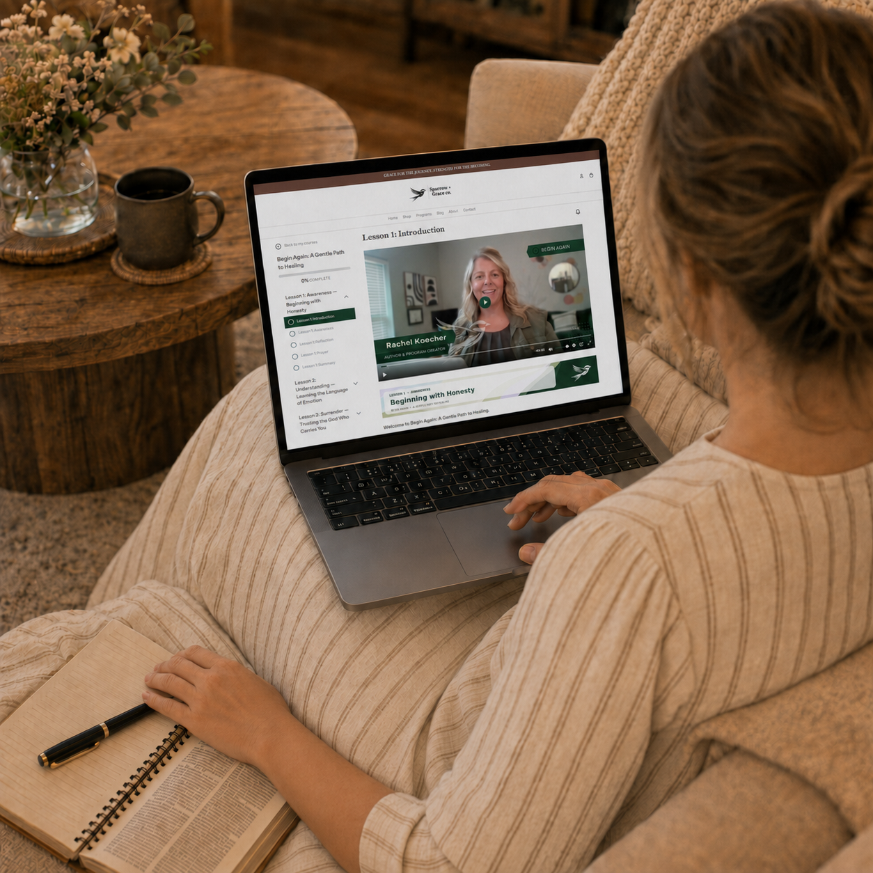 A woman sits on a beige couch in a cozy living room, viewed from behind, using a laptop to watch a video course. She is wearing a beige ribbed sweater and has an open notebook and pen on her lap.